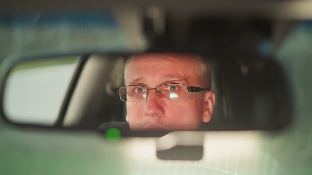 차의 뒷모습 거울에 반사되는 남자의 얼굴의 클로즈업, 그의 진지하고 집중된 표정을 포착합니다. 남자는 안경을 입고 있으며, 장면은 깊은 생각을 암시합니다.