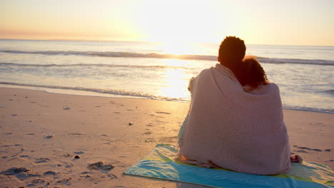 biracial couple wrapped in a blanket, watching a beach sunset with copy space