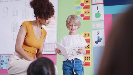 male pupil in elementary school classroom reading book to class with teacher