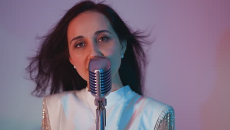 close-up of a singer in a white gown with beaded fringe, passionately singing as her hair flows gently against a pink background. the vintage microphone adds a classic touch