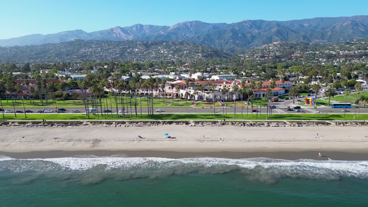 해안을 따라 바다 위를 날아다니는 아름다운 드론 영상, 해변에 부히는 파도, 멀리 있는 산, 종려나무, 해가 지면 이동하는 자동차를 보여준다.