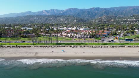 Hermosas-Imágenes-De-Drones-Volando-Sobre-El-Océano-A-Lo-Largo-De-La-Costa-Que-Muestran-Olas-Rompiendo-Contra-La-Orilla,-Montañas-En-La-Distancia,-Palmeras-Y-Automóviles-Viajando-Al-Atardecer