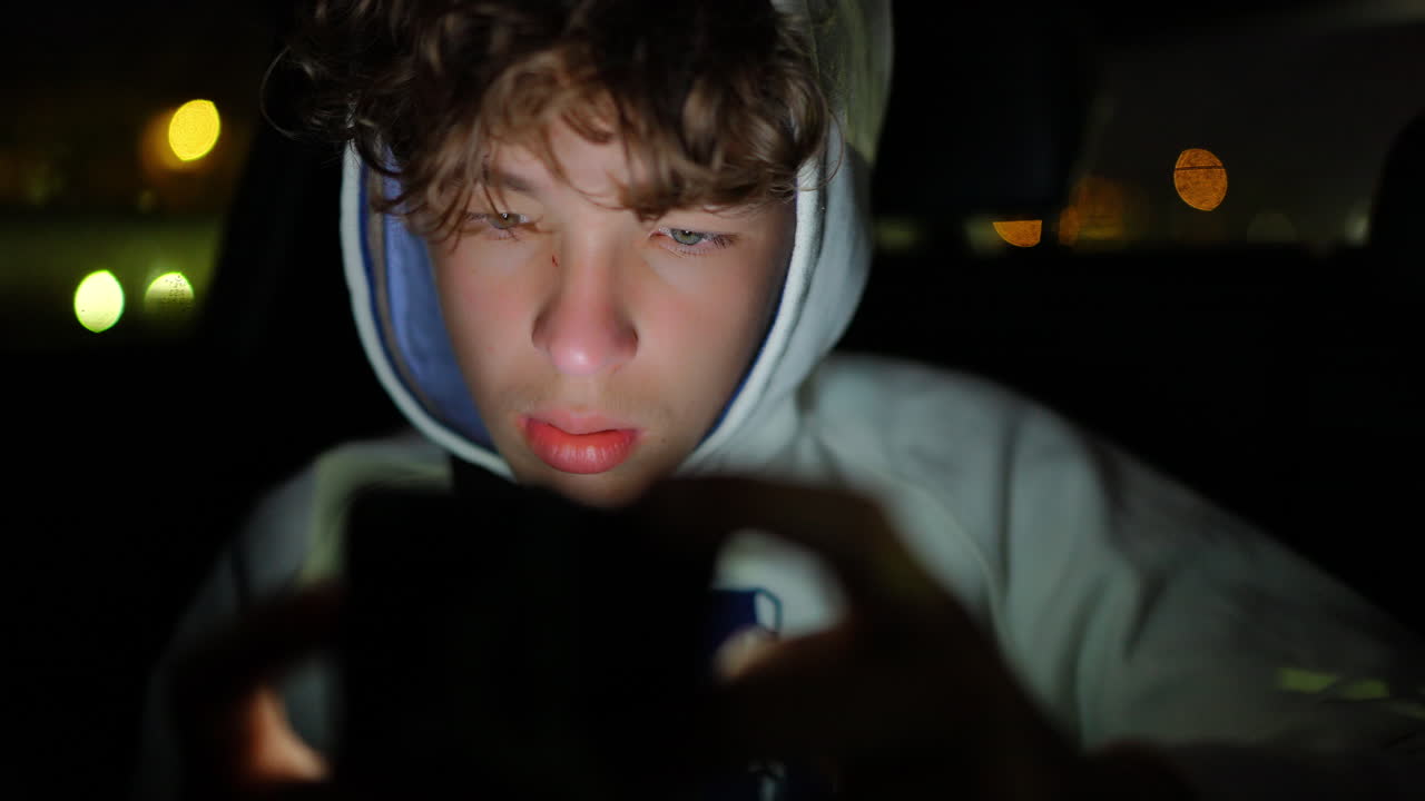 Teenage male passenger inside car during nighttime, immersed in smartphone screen, creating soft city lights bokeh in darkened background, capturing contemporary digital lifestyle moment