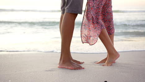 barefoot biracial couple stands on a sandy beach at sunset