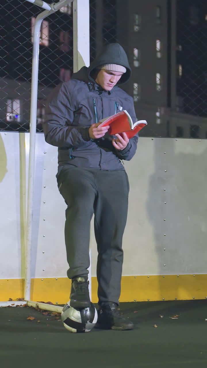 메탈 골포스트에 기대어 밤에 야외에서 책을 읽고, 발 에 축구공을 고, 도시 불빛으로 조명된 장면