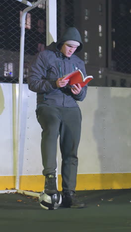 hombre con capucha y gorra apoyado contra un poste de meta metálico leyendo un libro al aire libre por la noche, una pelota de fútbol bajo los pies, una escena iluminada por las luces de la ciudad urbana