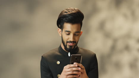 young cheerful indian man with beard and in black jacket smiling while texting on smartphone