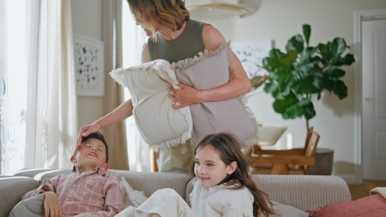 Carefree kids fighting pillows at sofa closeup. Mother calming naughty children