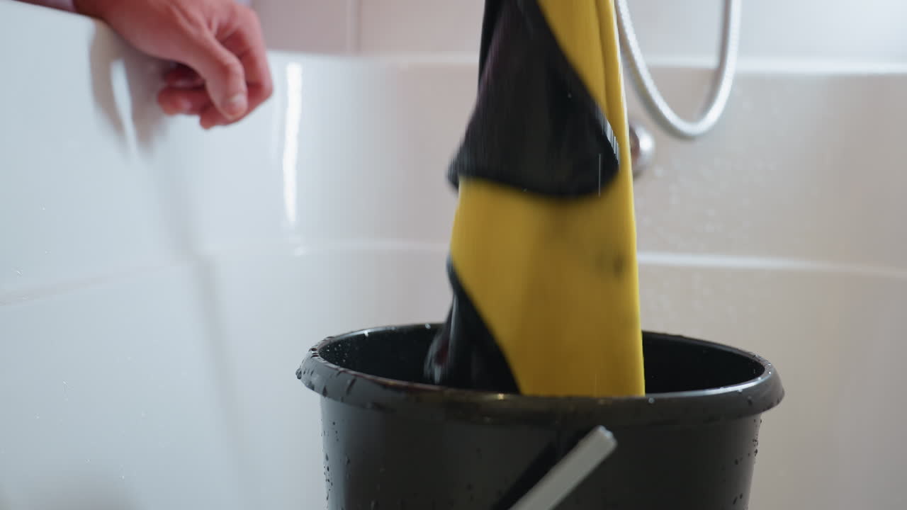 Close up of hand lifting yellow cloth from black bucket with streams of water dripping, fabric dripping liquid into container, scene of washing, cleaning, soaking and household task in progress