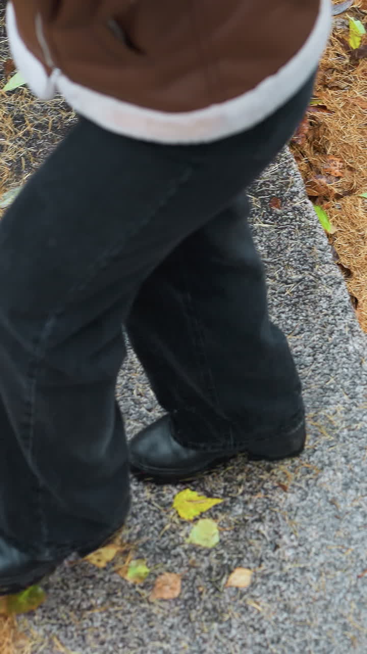A female walking in light snowfall, wearing brown shearling jacket, black trousers, walking on stone steps covered with colorful autumn leaves, face not visible