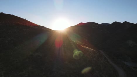 4k aerial of a drone ascending in front of dark mountains revealing the sun and lens flare with a light blue sky at sunset