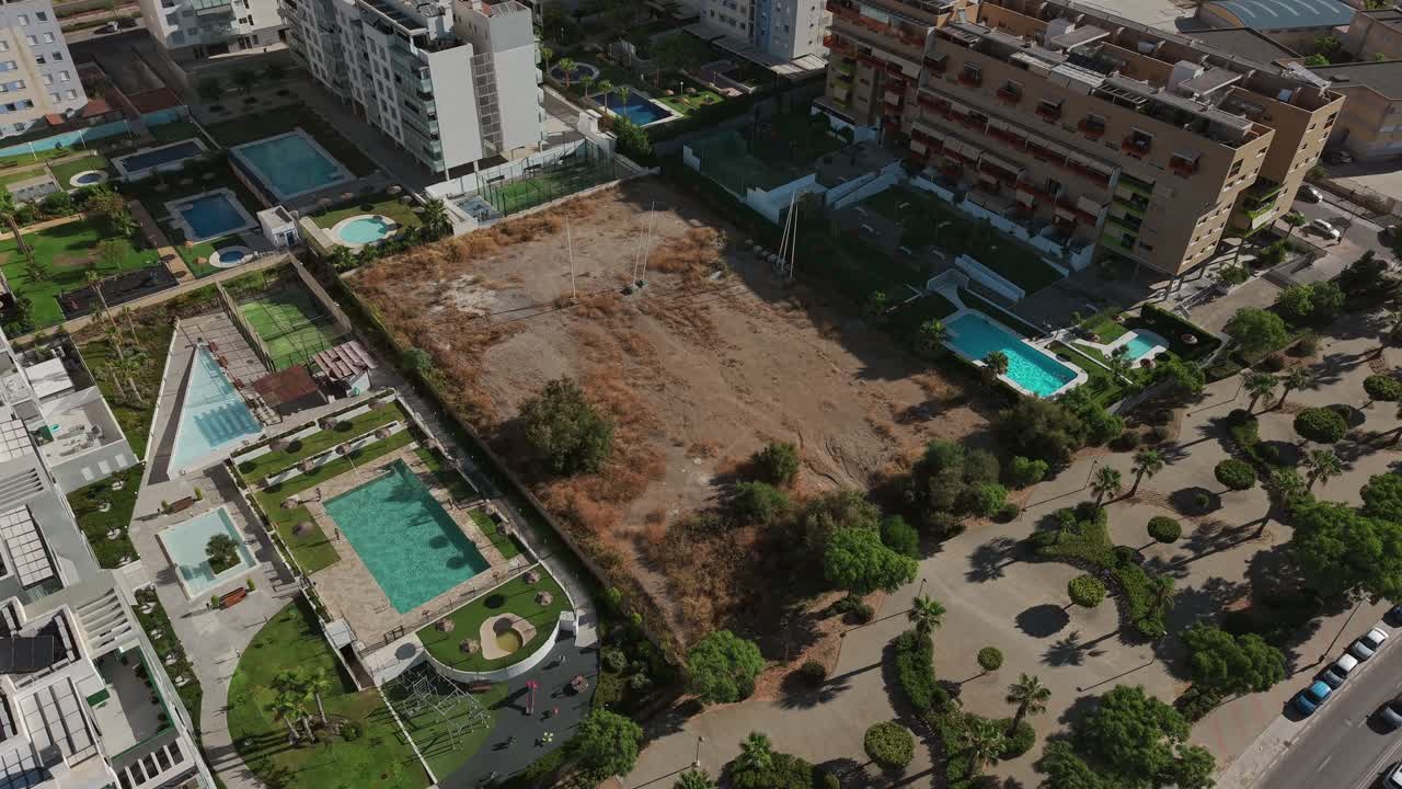 Aerial view of a vacant lot surrounded by modern apartment buildings with pools and landscaped paths