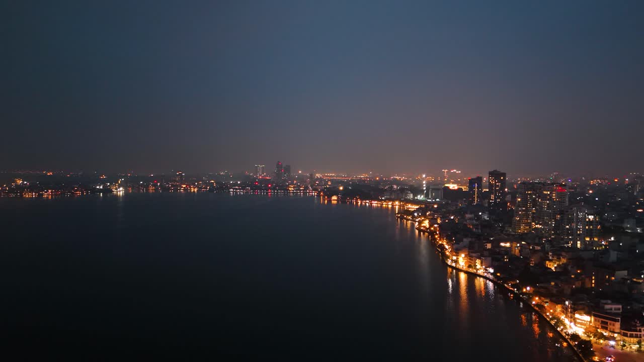 Drone footage of Hanoi's West Lake (Tay Ho) and the surrounding cityscape at night. City lights reflecting on the calm water, highlighting the vibrant urban shoreline. UHD