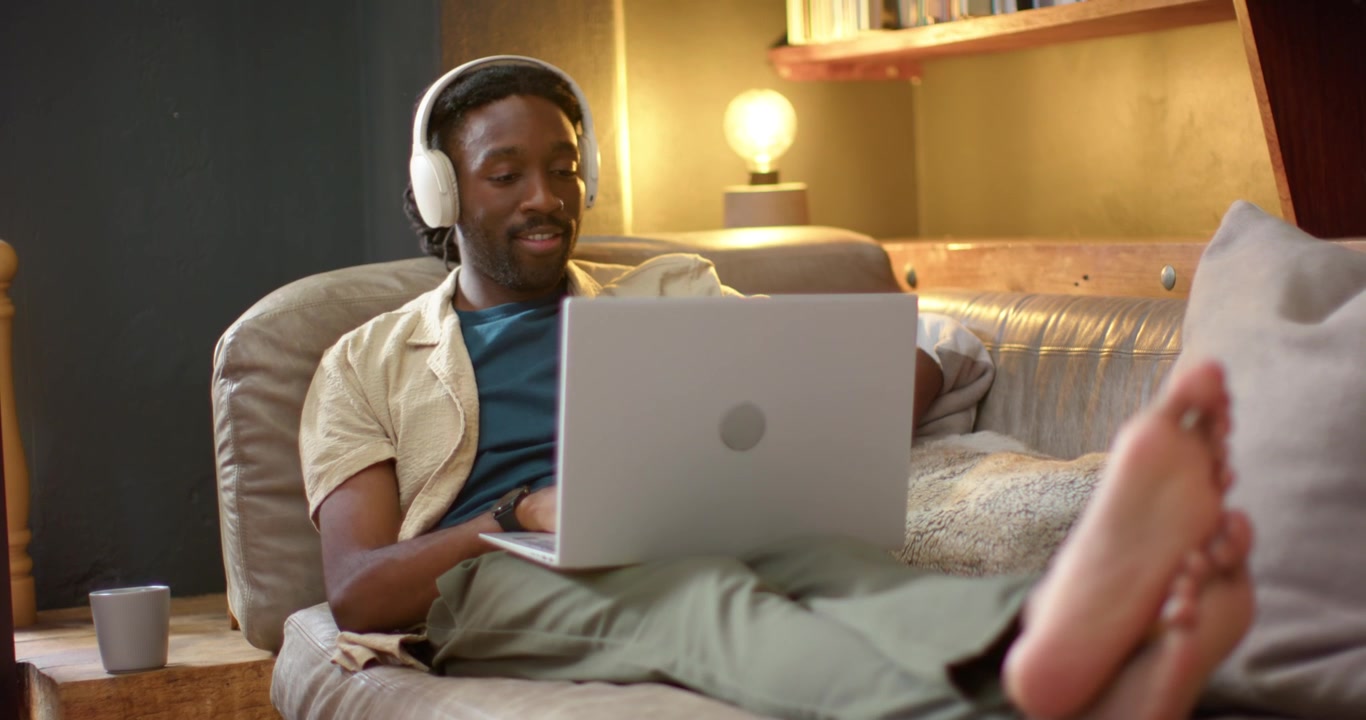 African American man lounging and typing on laptop on sofa for work, camera panning revealing lamp