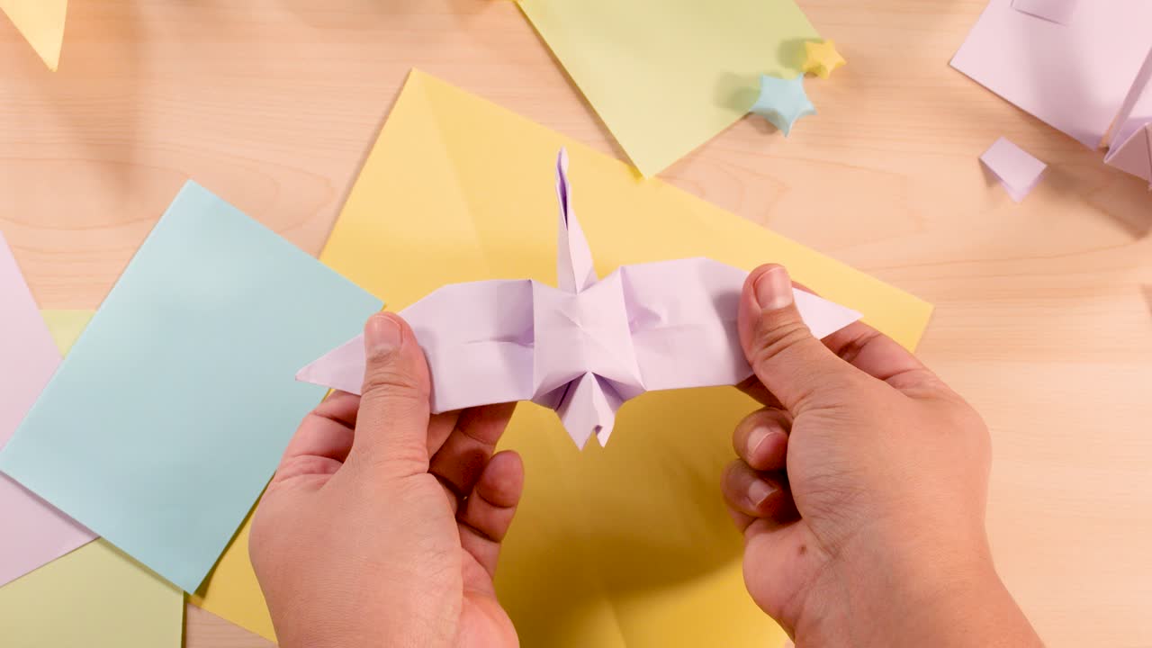 Hands gently flap origami bird wings on pastel paper, under soft, even lighting, overhead view