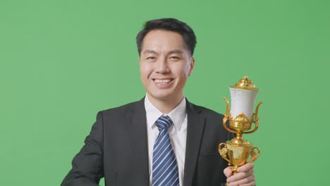 close up of asian business man in a suit and tie with a gold medal and trophy touching his chest then smiling and pointing to camera being proud of himself on green screen background in the studio