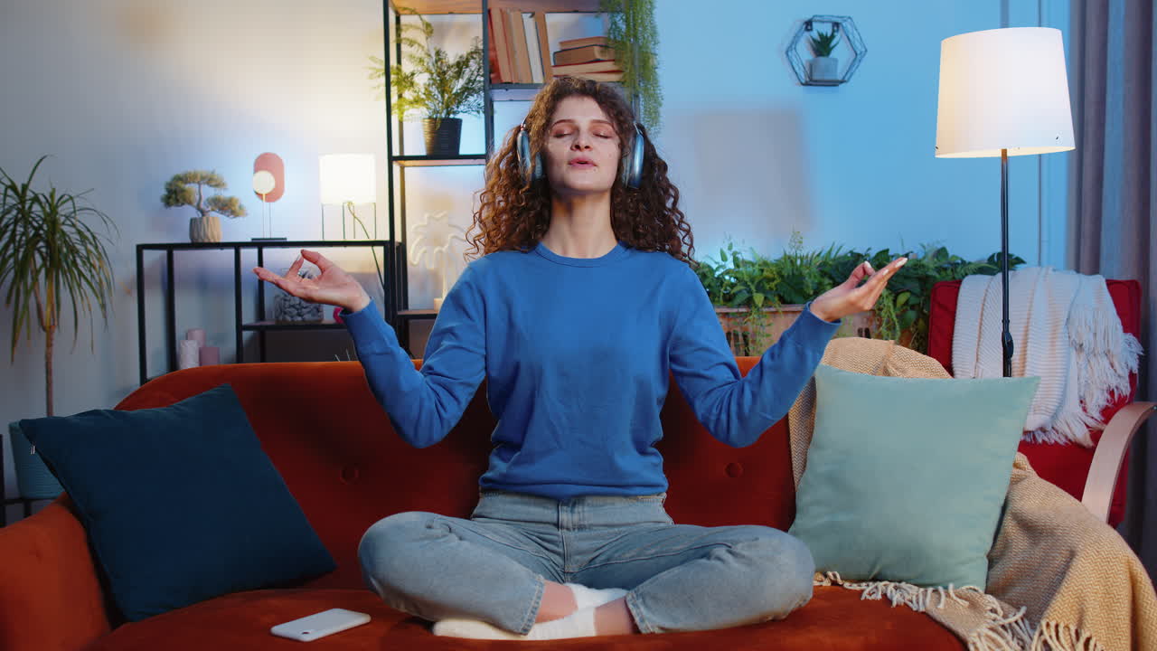 Relaxed young woman in headphones meditating in mudra gesture sitting on home sofa listening music