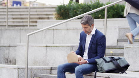 hombre de negocios sentado afuera en la pausa para el almuerzo comiendo comida para llevar de una caja de cartón reciclable sostenible