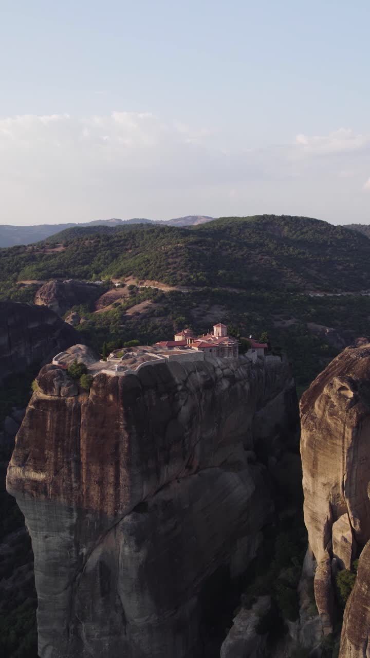 Vertical aerial rightward pan capturing the monastery’s elevated location with panoramic views of the Meteora landscape
