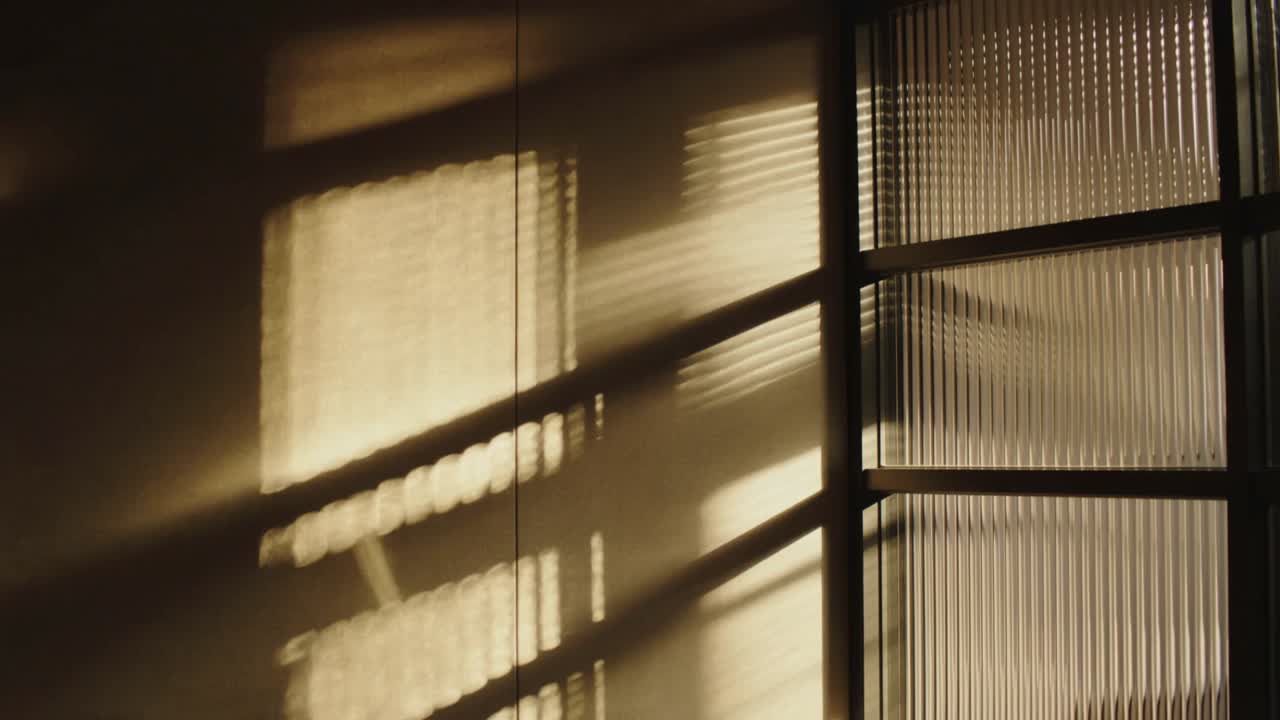 Luz del sol cambiante que se filtra por las persianas y crea bandas de sombra diagonales en la pared y el vidrio acanalado