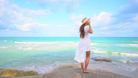 Mujer-Exótica-Sexy-Solitaria-En-Un-Vestido-Blanco-De-Pie-Sobre-Una-Roca-Frente-A-Las-Olas-Del-Mar-Tropical-Con-Sombrero-De-Verano-Disquete,-Cámara-Lenta-De-Fotograma-Completo