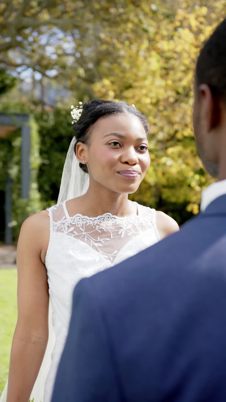 Vertical video of african american couple smiling at wedding ceremony in garden, in slow motion