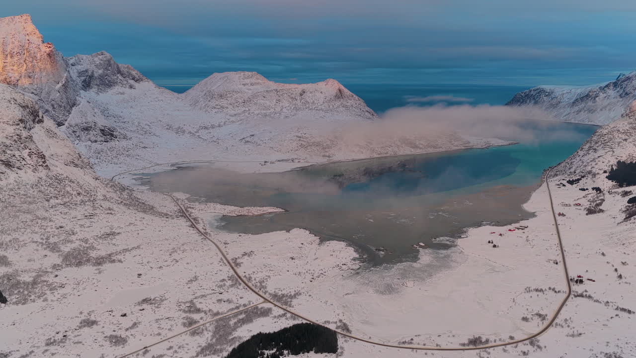 Norveç'teki karla kaplı Lofoten adalarının nefes kesen drone çekimi, altın saatte virajlı bir yol, turkuaz su ve alçak bulutları gösteriyor.