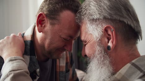 Close-up-an-elderly-man-with-gray-hair-a-lush-beard-and-an-earring-in-his-ear-in-a-checkered-shirt-presses-his-forehead-to-his-brunette-boyfriend-in-a-checkered-shirt-and-holds-him-by-the-shoulder-in-a-modern-apartment-of-an-LGBT-couple