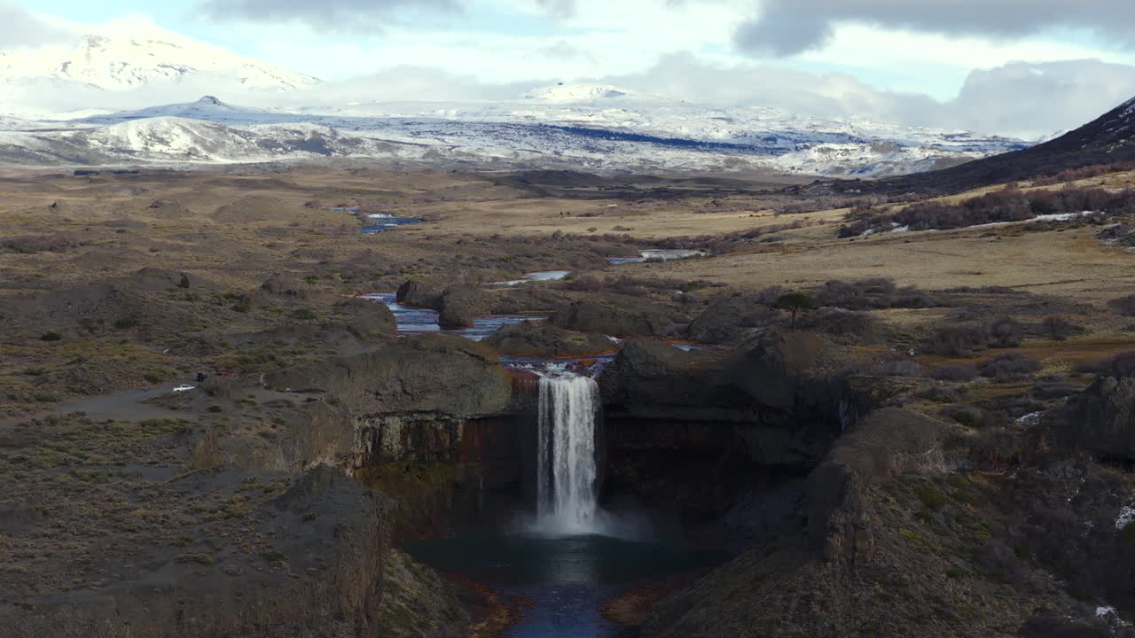 Aerial shot descending toward Salto del Agrio waterfall with surrounding volcanic rocks and snowy Andes mountains in Caviahue, Argentina