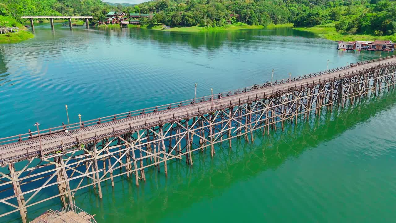 Aerial orbit around the Mon Bridge in Sangkhlaburi, Thailand. The long wooden bridge spans calm blue water with reflections, connecting the village and floating houses