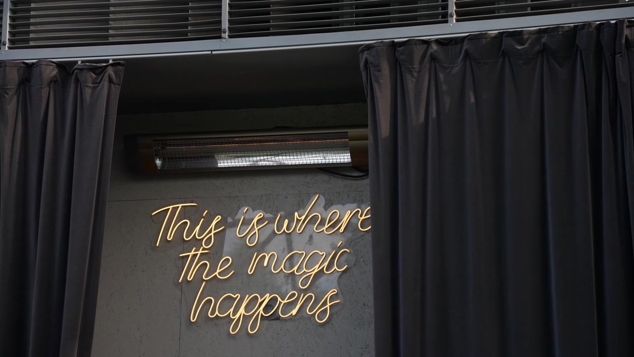 Insegna al neon luminosa con la scritta 'Qui è dove accade la magia' risplende su una parete grigia ed elegante, incorniciata da tende nere. Illuminazione interna soffusa, telecamera fissa, composizione cinematografica.