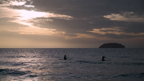 silhouetted two women paddleboarding at sunset on their sups with sulug islet in background, in malaysia sabah