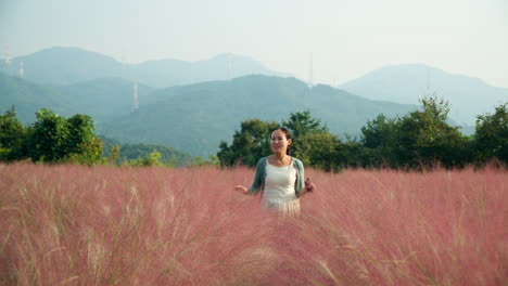 asian korean woman walks towards camera through pink muhly tall grass field in slow motion with scenic mountain range background in pocheon highlands herb island farm