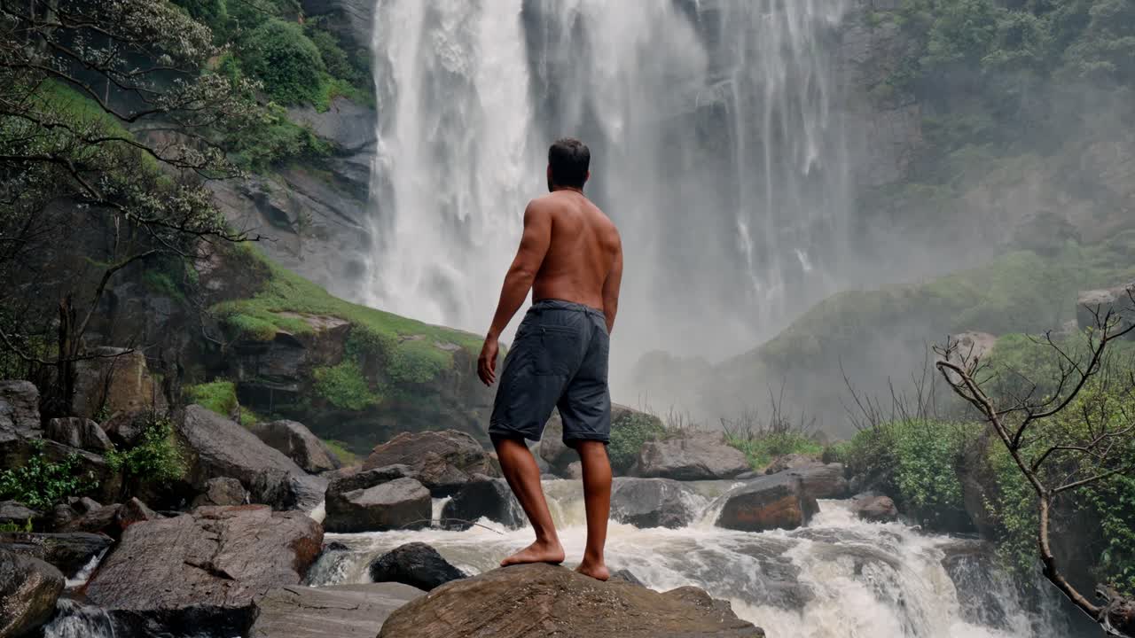 A lone man stands in front of the powerful Bomburu Ella Waterfalls, surrounded by lush forest and rocky terrain in the misty highlands of Nuwara Eliya, Sri Lanka.