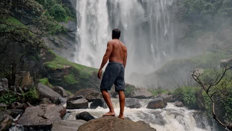 a lone man stands in front of the powerful bomburu ella waterfalls, surrounded by lush forest and rocky terrain in the misty highlands of nuwara eliya, sri lanka.