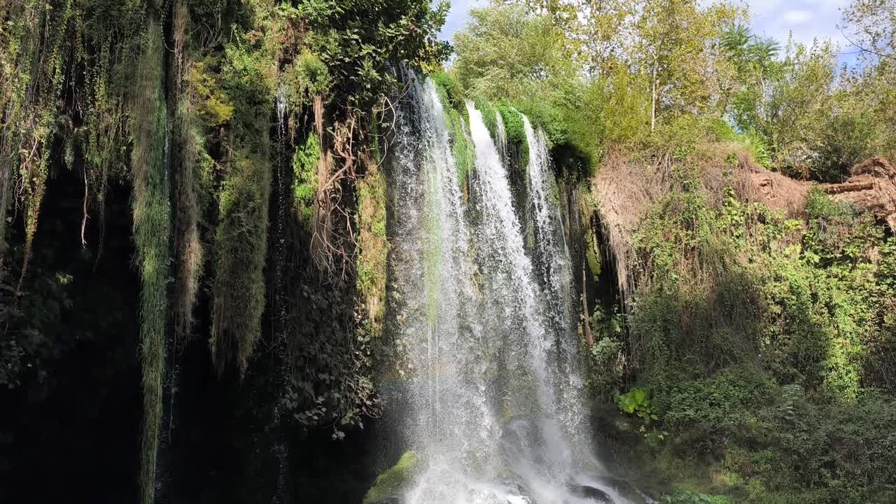 Camera captures Duden Waterfall's powerful flow from below, ascending perspective revealing water curtain against sky. Highlights mist dynamics and rock formations. Ideal for nature documentaries