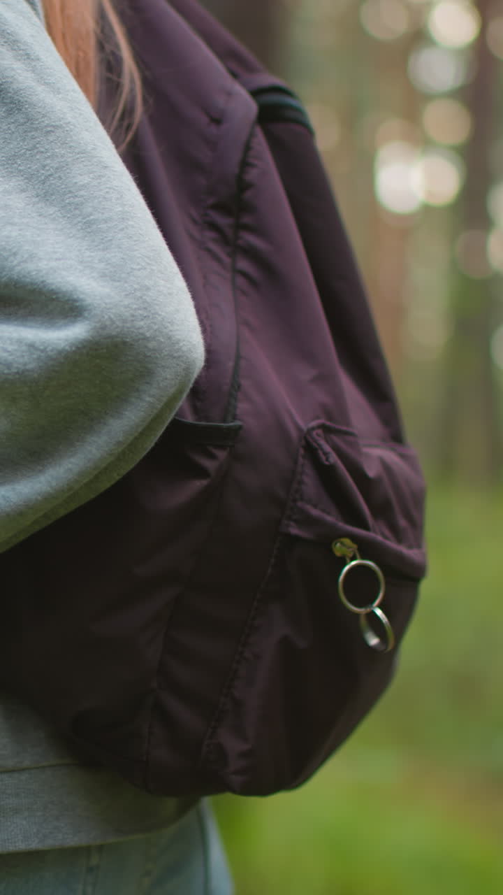 Close-up of young woman hiking through forest wearing gray sweater and backpack, standing amidst lush greenery, focus on arm and hand grasping backpack strap