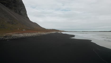 Luftflug-Entlang-Des-Schwarzen-Sandstrandes-Stokksnes,-Vulkanische-Dunkle-Berge-In-Der-Ferne,-Düstere,-Stimmungsvolle,-Wolkige-Landschaft-Islands