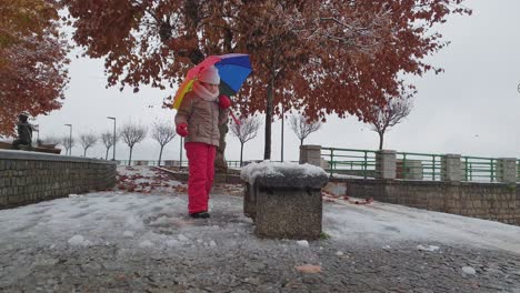 child girl with multicolored umbrella and red trousers plays kicking snow