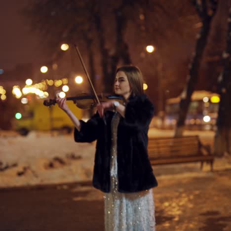 a gorgeous girl in a bright dress and black coat performs classical music on a violin in a winter evening in the centre of the town. blurred background. camera motion to left