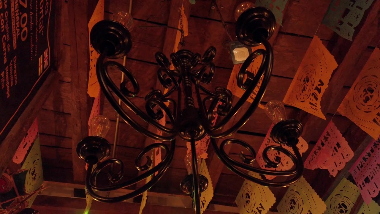 Vista dal basso di un lampadario decorativo in ferro battuto sotto un soffitto in legno. La luce calda illumina striscioni colorati di papel picado, mettendo in risalto l'arredamento interno tradizionale e la dettagliata lavorazione del metallo.