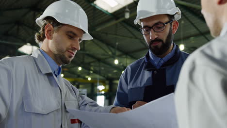 close-up view of three engineers wearing helmets and holding blueprint while talking in a factory