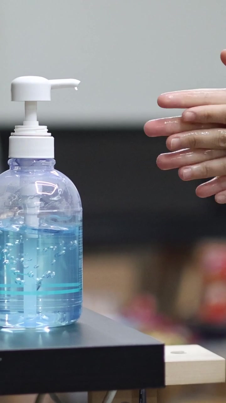 Una toma vertical que muestra una botella de gel desinfectante azul mientras unas manos se frotan cerca.