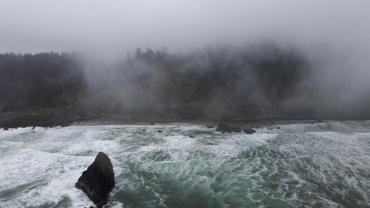 White foamy waves crash on a fog shrouded rugged ocean beach, aerial track