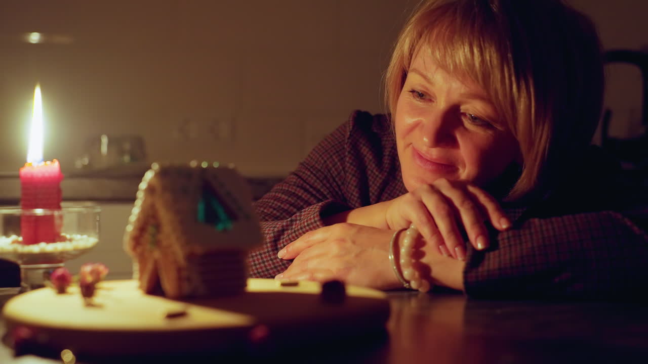 Thoughtful woman rests hands on table, gazing at intricately decorated gingerbread house, warm candlelight casts a soft glow, creating a cozy and contemplative atmosphere