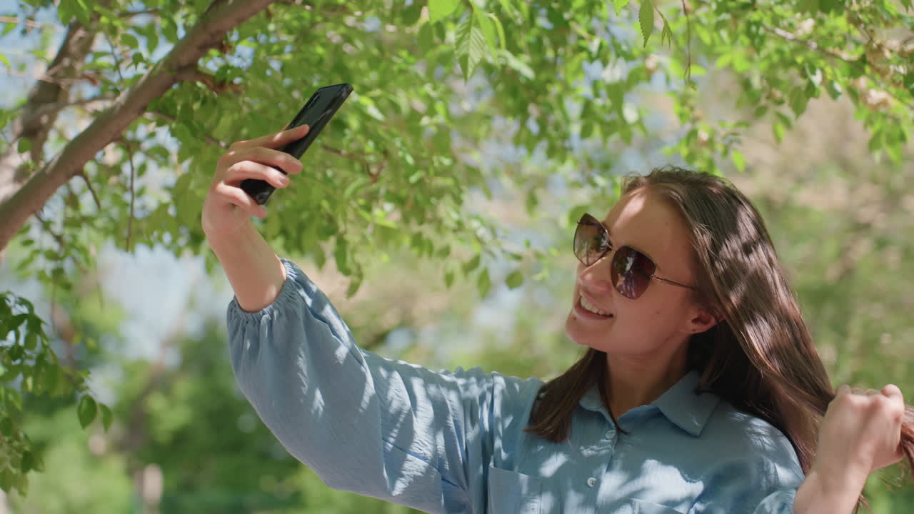 Ella captura momentos, ejercicios individuales de técnicas para selfis en exteriores, persona practicando selfis bajo la cubierta de un árbol frondoso, una mujer creativa se ajusta el pelo y el teléfono al aire libre, a la luz del día.
