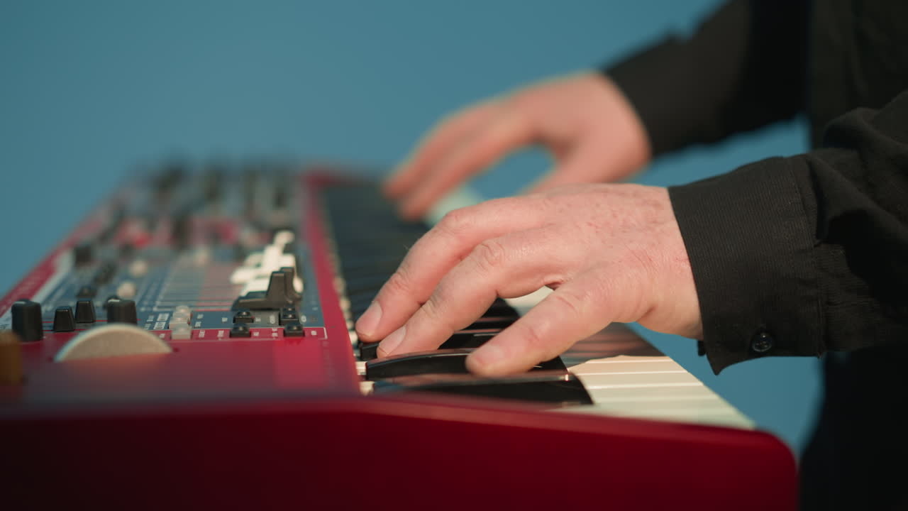 얼굴이 없는 키보디스트가 파란색 배경에 빨간색 플러 피아노를 연주하고 있다. 클로즈업 은 음악가의 손이 키를 고 있는 것을 강조한다.