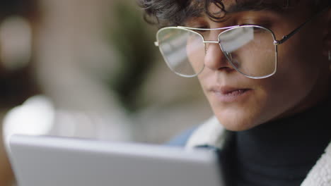 Cerrar-Un-Atractivo-Joven-Estudiante-Usando-Una-Tableta-En-Un-Café-Navegando-En-Línea-Leyendo-Mensajes-De-Redes-Sociales-Disfrutando-Bebiendo-Café-Relajándose-Usando-Gafas-Elegantes