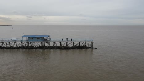 Vista-Aérea-De-Personas-En-Un-Largo-Muelle-Sobre-El-Agua-En-El-Vasto-Océano-Sin-Fin