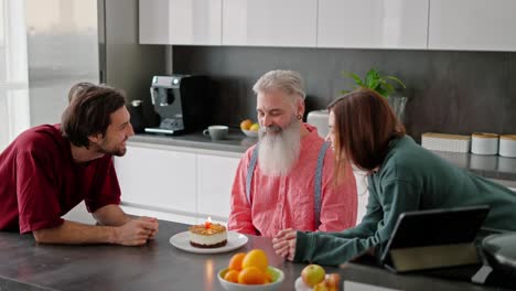 A-happy-brunette-man-with-stubble-in-a-red-T-shirt-together-with-his-brunette-sister-in-a-green-sweater-congratulate-their-elderly-dad-with-a-lush-beard-in-a-pink-shirt-on-his-birthday-and-together-blow-out-a-candle-on-a-small-cake-while-sitting-in-a-modern-kitchen-in-an-apartment